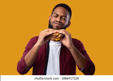 Happy African Man Eating Burger Posing Standing Over Yellow Studio Background. Black Guy Tasting Cheeseburger Enjoying Unhealthy Junk Food. Nutrition And Overeating Habit