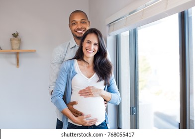 Happy african man embracing his pregnant girlfriend and looking at camera. Loving husband gently hugging pregnant wife at home. Multiethnic couple expecting a baby.