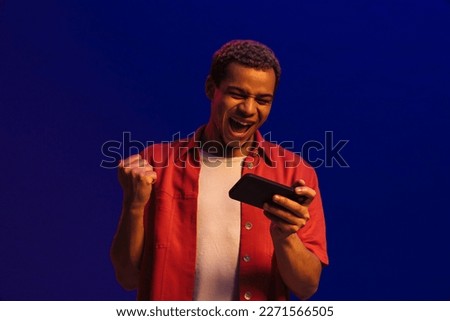 Happy african man gesturing as winner while playing online game on mobile phone isolated over blue neon studio wall