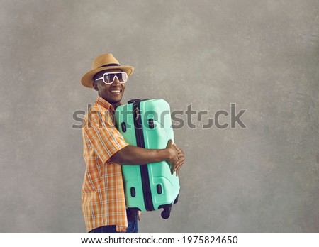 Happy African man going on summer vacation. Portrait of cheerful black tourist in sunglasses, sun hat and orange shirt smiling and holding mint green travel suitcase standing on gray studio background