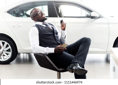 Happy African Man With Keys In His Hands Happy Buying A New Car Sitting On A Chair In The Showroom