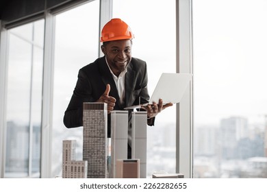 Happy african man in orange construction hard hat and suit showing thumb up and smiling on camera. Male inspector looks satisfied with table with skyscraper building maquette.