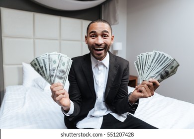 Happy African man in suit with money in hands sitting on bed with open mouth in hotel room Stock Photo