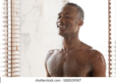 Happy african man taking cold shower refreshing in the morning, washing body standing under falling water In modern bathroom at home. Wellness, hygiene and male bodycare routine. Empty space