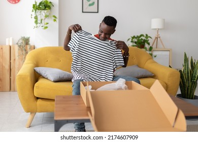 Happy African Man Unboxing Package After Delivery - Young Adult Male Holding Striped Shirt Sitting On Sofa At Home - American Customer Receiving Cardboard Parcel Box - Online Shopping Concept