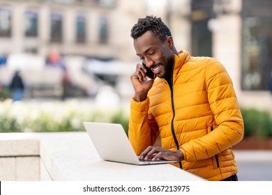 Happy african man using laptop and talking on smartphone on balcony in city