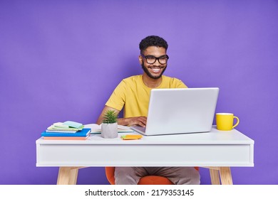 Happy African man using laptop while sitting at the desk against purple background - Shutterstock ID 2179353145