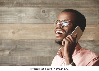 Happy African man wearing glasses, talking on mobile phone with his family while being away on business trip, posing against wooden copy space wall for your information. Technology and connection