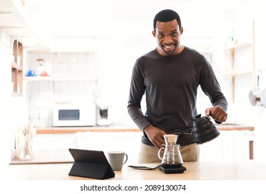 Happy African middle age man learning online making coffee class via digital tablet and looking at camera, poured hot water from the kettle into the glass jar to make coffee in modern kitchen at home. - Powered by Shutterstock