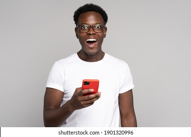Happy African Millennial Man In Glasses Wear White T-shirt, Holding Mobile Phone, Isolated On Studio Grey Background. Black Guy Smiling Broadly, Using Smartphone In Red Case, Looking At Camera.