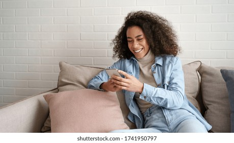 Happy African Millennial Woman Holding Smart Phone Watching Social Media Video Content. Smiling Young Black Teen Gen Z Girl Using Funny Mobile App Laughing, Playing Game, Chatting On Cell At Home.