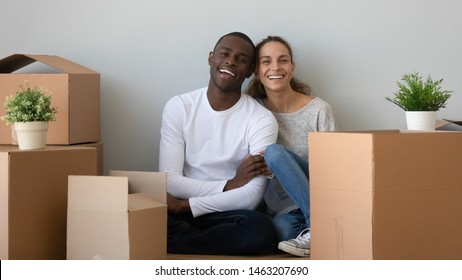 Happy african mixed race ethnicity couple first time home buyers looking at camera sit on floor in new home with boxes, smiling man and woman on moving day, relocation, mortgage concept, portrait