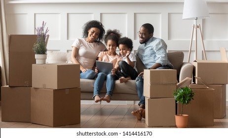 Happy African Parents And Cute Little Children Using Digital Tablet On Moving Day Sit On Sofa With Boxes In Living Room, Black Family With Kids Hold Computer Having Fun Renovate Relocate In New Home