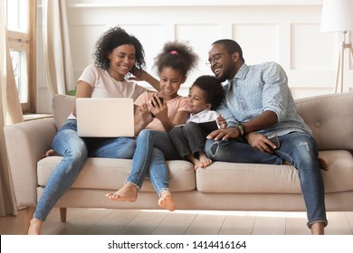 Happy african parents and kids obsessed addicted to gadgets use laptops digital tablet phone sit on sofa, black family mom dad and children have fun with devices at home internet technology addiction