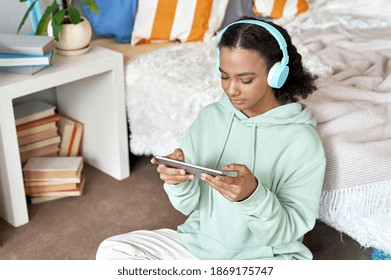 Happy african teen girl wearing headphones calling, watching videos using digital tablet at home. Mixed race teenager having online virtual chat videocall using computer sitting in bedroom on floor. - Powered by Shutterstock