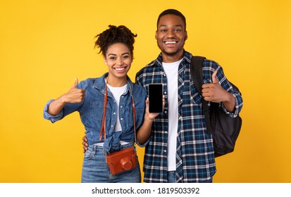Happy African Travellers Couple Showing Smartphone Empty Screen Gesturing Thumbs Up, Recommending Mobile Travel App Standing On Yellow Studio Background. Approving Application For Tourists Concept