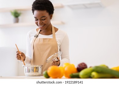 Happy African Woman Cooking Dinner In A Pot Standing In Modern Kitchen At Home, Wearing Apron. Black Housewife Preparing Meal Indoors. Healthy Food Recipes, Household And Nutrition