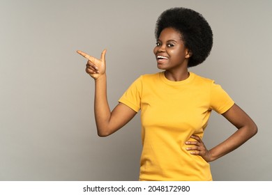Happy african woman point finger left with excited cheerful smile. Young black girl with afro hairstyle present show new product or sale advertising space isolated over gray studio wall background