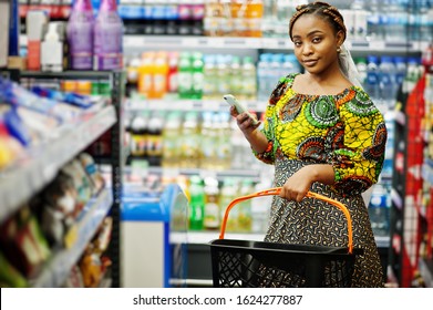 Happy African Woman In Traditional Clothes And Veil Looking Product At Grocery Store, Shopping In Supermarket.  Afro Black Women With Mobile Phone.