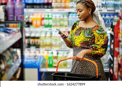 Happy African Woman In Traditional Clothes And Veil Looking Product At Grocery Store, Shopping In Supermarket.  Afro Black Women With Mobile Phone.