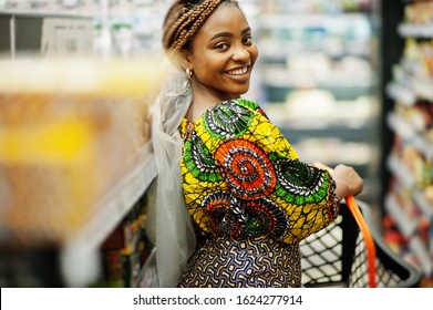 Happy African Woman In Traditional Clothes And Veil Looking Product At Grocery Store, Shopping In Supermarket.  Afro Black Women Costumer With Basket Buying Food At The Market.