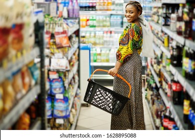 Happy African Woman In Traditional Clothes And Veil Looking Product At Grocery Store, Shopping In Supermarket.  Afro Black Women Costumer With Basket Buying Food At The Market.