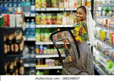 Happy African Woman In Traditional Clothes And Veil Looking Product At Grocery Store, Shopping In Supermarket.  Afro Black Women Costumer With Basket Buying Food At The Market.