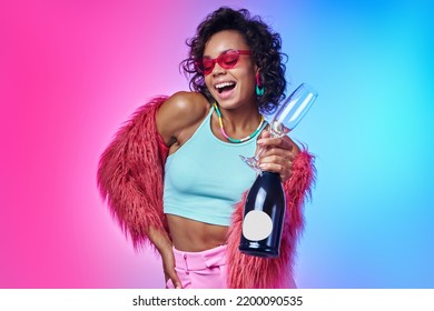 Happy African woman in trendy clothes holding bottle and sparkling wine against colorful background