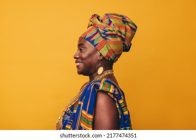 Happy African woman wearing a traditional attire against a yellow background. Mature black woman dressed in colourful Kente cloth and golden jewellery. Smiling woman embracing her rich culture.