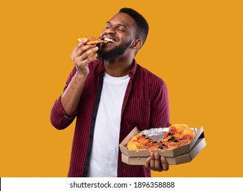 Happy African Young Man Enjoying Pizza Biting Tasty Slice Posing With Box Over Yellow Background. Junk Food Lover Eating Italian Pizza In Studio. Unhealthy Male Nutrition And Cheat Meal
