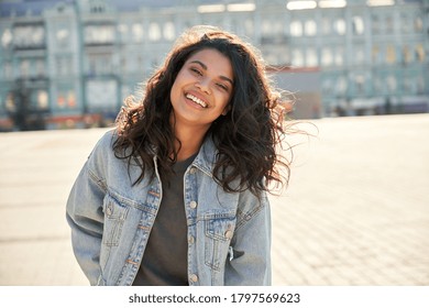 Happy African young woman wearing denim jacket laughing looking at camera standing on street. Smiling Afro American teen generation z hipster girl posing outdoor backlit with sunlight, portrait.
