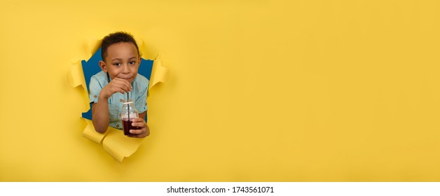 Happy African-American Boy In Blue Shirt Is Holding Juice Bottle, Drinking Red Cherry Drink From Black Bar Straw, Against Yellow Background Of Torn Paper Wall. Healthy Drink Detoxification Concept.