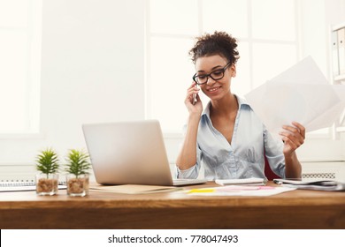 Happy african-american business woman talking on mobile at office. Young female manager consulting on phone, holding papers, using laptop at working place, copy space - Shutterstock ID 778047493