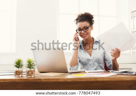 Happy african-american business woman talking on mobile at office. Young female manager consulting on phone, holding papers, using laptop at working place, copy space