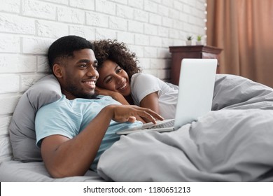 Happy african-american couple in bed with laptop browsing in internet, copy space