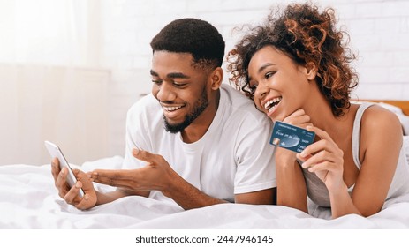 Happy african-american couple buying online, using smartphone and credit card in bed, enjoying shopping in internet, free space - Powered by Shutterstock