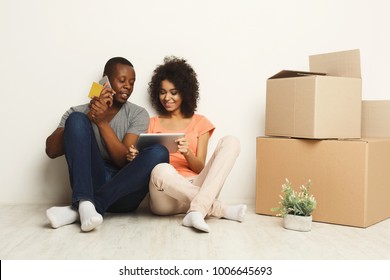 Happy african-american couple discussing design of new appartment, choosing color scheme on tablet, making order on mobile, sitting on floor at white wall near moving and storage boxes, copy space - Powered by Shutterstock