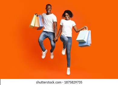 Happy African-american Couple With Purchases Holding Hands And Jumping Up, Smiling At Camera, Orange Studio Background. Joyful Black Man And Woman Showing Bunch Of Shopping Bags. Sale, Discount, Deal