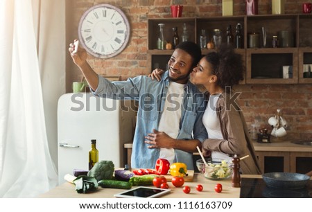 Happy african-american couple taking selfie with smartphone while preparing salad at kitchen