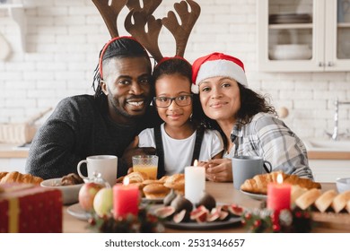 Happy african-american family, parents and small daughter in Santa Claus hat celebrating Christmas and New Year together at home, looking at camera during holiday breakfast with presents gifts - Powered by Shutterstock