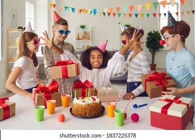 Happy African-American girl receiving birthday presents sitting at festive table with cake. Group of children in funny hats and sunglasses giving presents to their friend during fun party at home