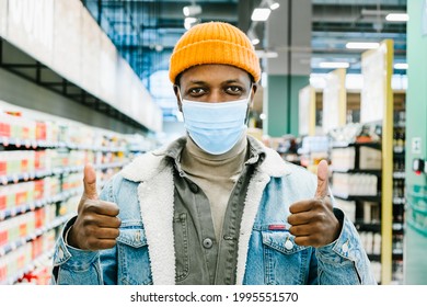 Happy African-American guy in knitted hat puts and protective mask shows thumbs up standing in supermarket department closeup
