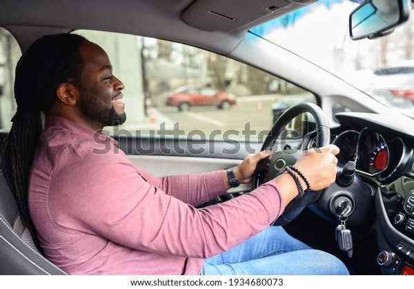 A happy African-American man with the dreadlocks is\
driving a car, side view from a passenger seat on a cheerful\
multiracial man holding a steering wheel, bought a car, learned to\
drive