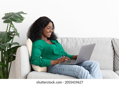 Happy African-american Millennial Lady Freelancer Working Online From Home, Reclining On Couch By Empty White Wall At Living Room, Using Modern Laptop, Chatting With Guys, Copy Space