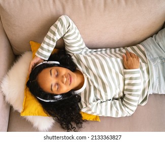 Happy african-american woman lying on sofa and listening to music in headphones. Enjoying audio playing in earphones. Relaxing and chilling at home. Positive emotions. Having fun while laying along
