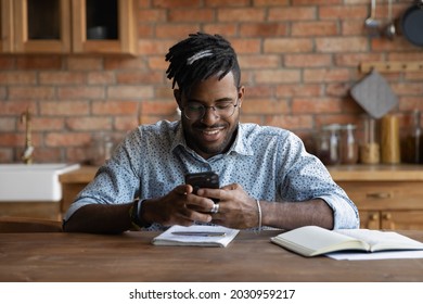 Happy Afro American Guy Chatting Online, Checking Messages, Reading, Texting, Smiling, Looking At Screen. Black Young Man Using Online App On Mobile Phone, Studying Or Working In Home Kitchen