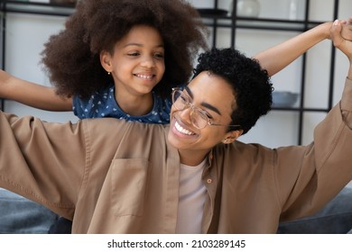 Happy Afro American Mom And Daughter Kid Playing Airplane At Home. Girl Leaning On Mums Back, Making Open Flying Hands, Smiling, Having Fun. Mom And Kid Affection, Family Relationship