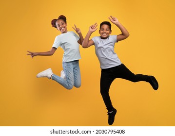 Happy afro american school-aged brother and sister fooling on yellow studio background, full length photo. Positive black boy and girl siblings jumping up, smiling and gesturing, having fun together