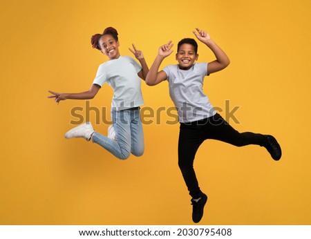 Happy afro american school-aged brother and sister fooling on yellow studio background, full length photo. Positive black boy and girl siblings jumping up, smiling and gesturing, having fun together