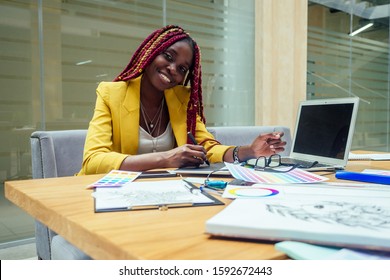 Happy, Afro American Woman In A Stylish Yellow Jacket And Multi-colored Dreadlocks Pigtails Graphic Designer Draws A Sketch On A Tablet Workplace In A Light Large Modern Office